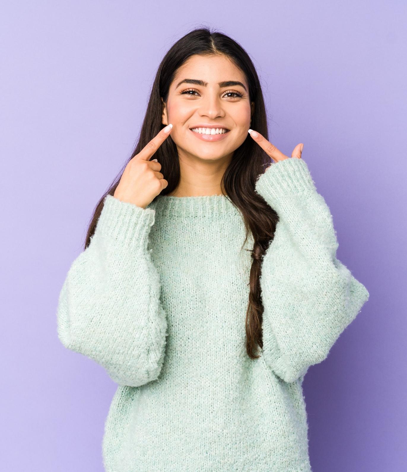 young indian woman isolated on purple background smiles pointing fingers at mouth